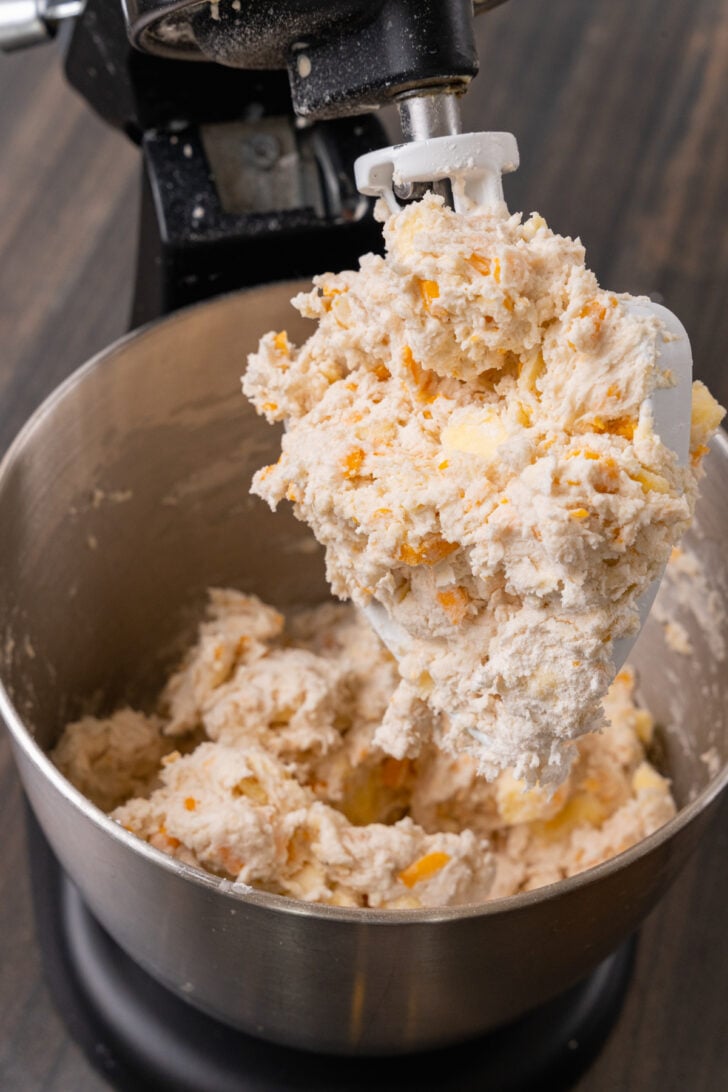 Metal mixing bowl on a black stand mixer with cheddar cheese biscuit dough on a metal mixing paddle.