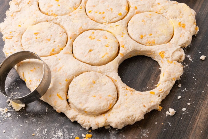 Cheddar cheese biscuit dough rolled out on a wooded board and cut with a metal round cutter.