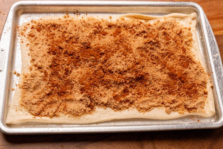 Raw pizza dough rolled out on a silver baking sheet topped with brown sugar and cinnamon.