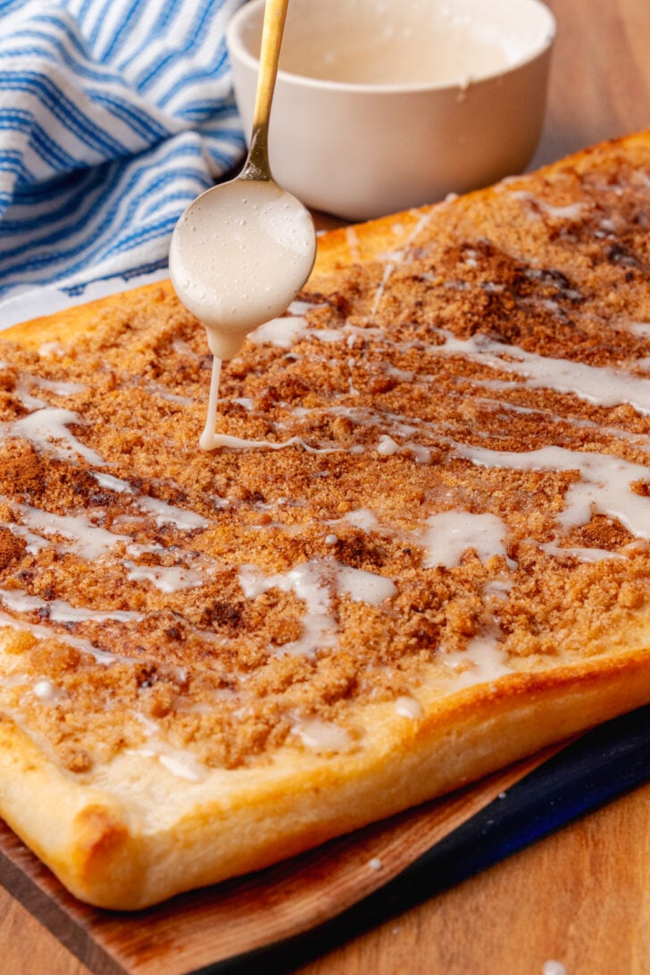 Baked cinnamon brown sugar pizza being topped with vanilla icing drizzled with a gold spoon.