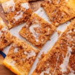 Squares of cinnamon brown sugar pizza topped with vanilla icing on a parchment paper on a wooden board.