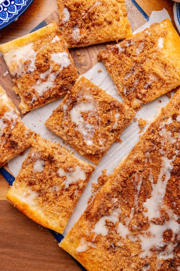 Squares of cinnamon brown sugar pizza topped with vanilla icing on a parchment paper on a wooden board.