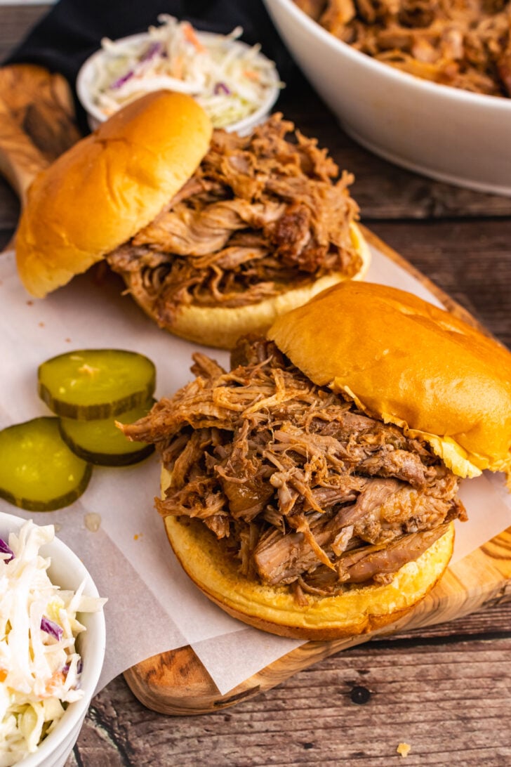 Pulled Pork piled on a round bun on a wooden cutting board served with sliced pickles and coleslaw.