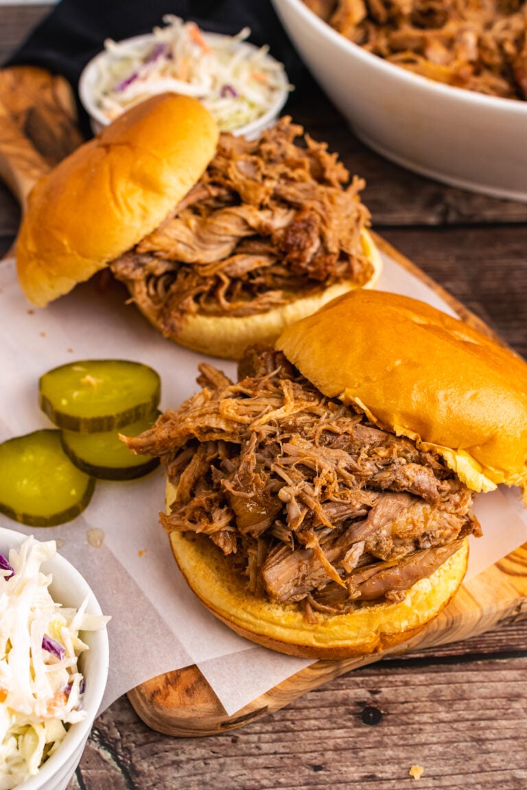 Pulled Pork piled on a round bun on a wooden cutting board served with sliced pickles and coleslaw.