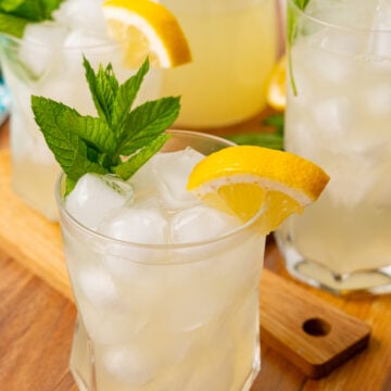 Glass of homemade lemonade with a lemon wedge and sprig of mint on a wooden board.