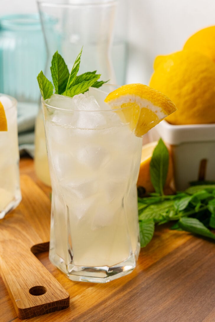 Glass of homemade lemonade with a lemon wedge and sprig of mint on a wooden board.