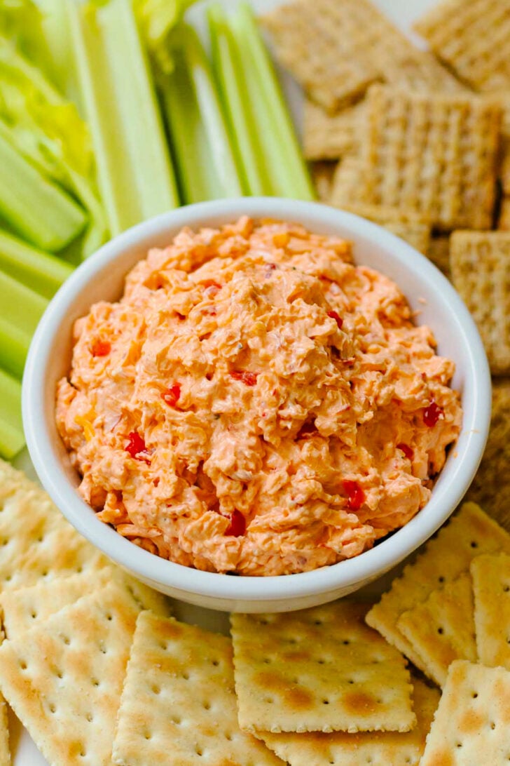 Homemade Pimento Cheese in a white bowl served and wooden knife with saltine crackers and celery sticks on a white platter.