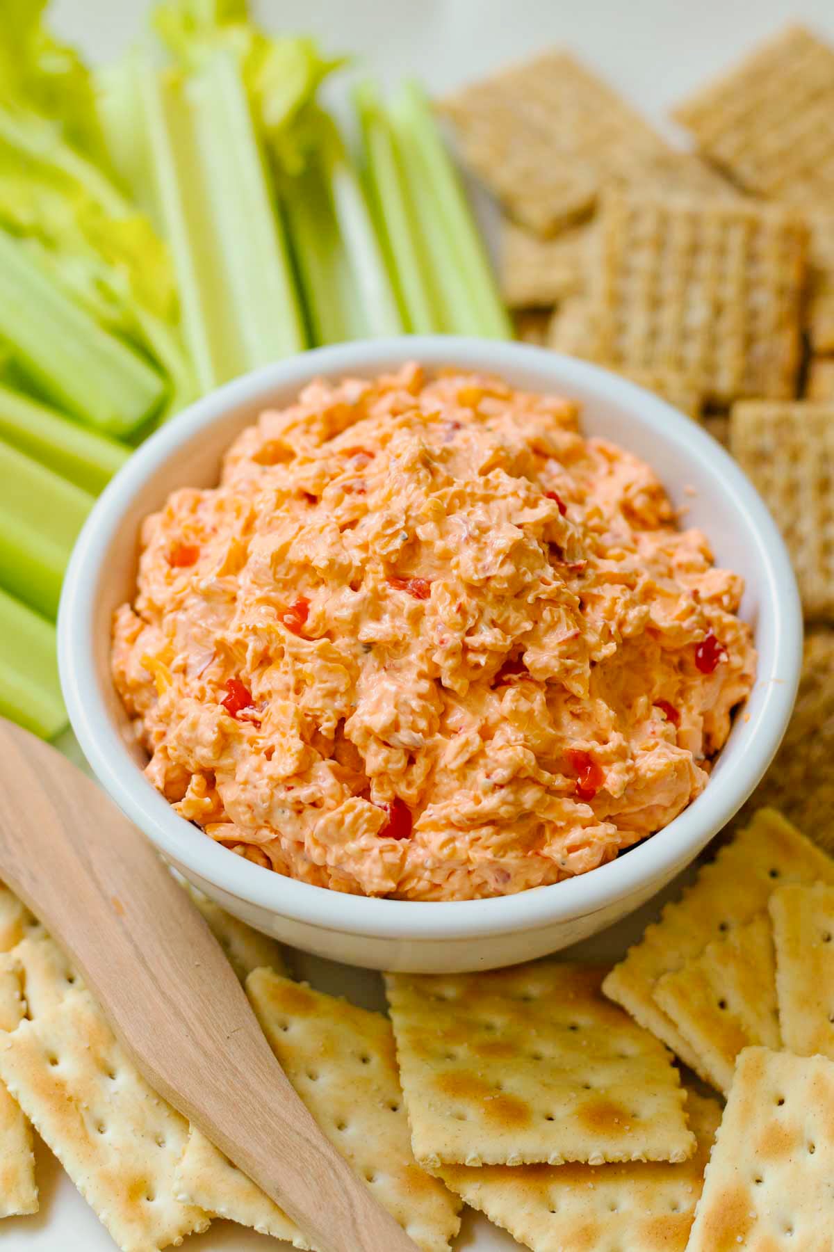 Homemade Pimento Cheese in a white bowl served and wooden knife with saltine crackers and celery sticks on a white platter.