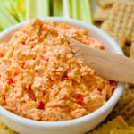 Homemade Pimento Cheese in a white bowl served and wooden knife with saltine crackers and celery sticks on a white platter.