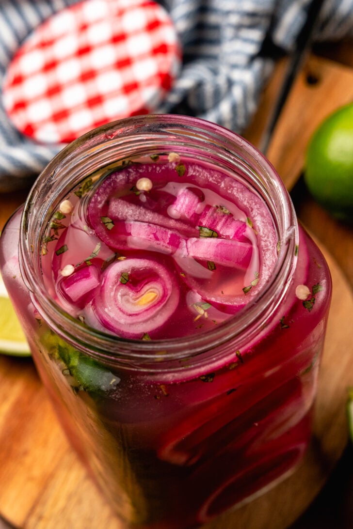 Large glass jar with red onions, sliced jalapeno pepper, garlic and parsley on a woodend board.