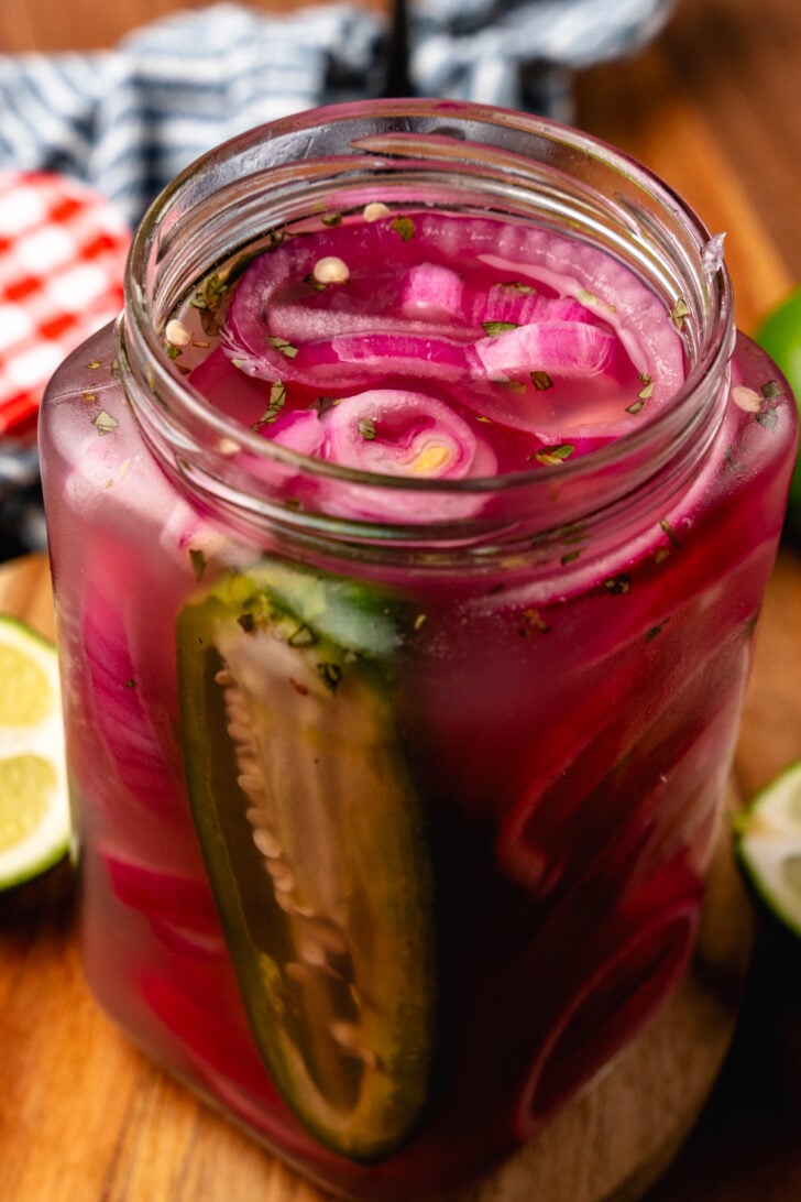 Large glass jar with red onions, sliced jalapeno pepper, garlic and parsley on a woodend board.