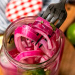 Pickled red onions on a fork pulled out of a far that have fresh parsley and jalapeno peppers in the jar.