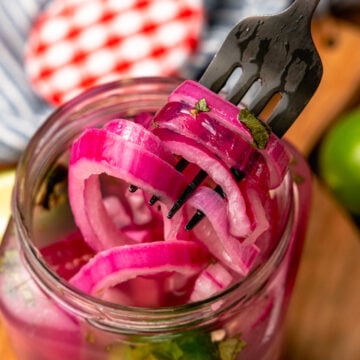 Pickled red onions on a fork pulled out of a far that have fresh parsley and jalapeno peppers in the jar.