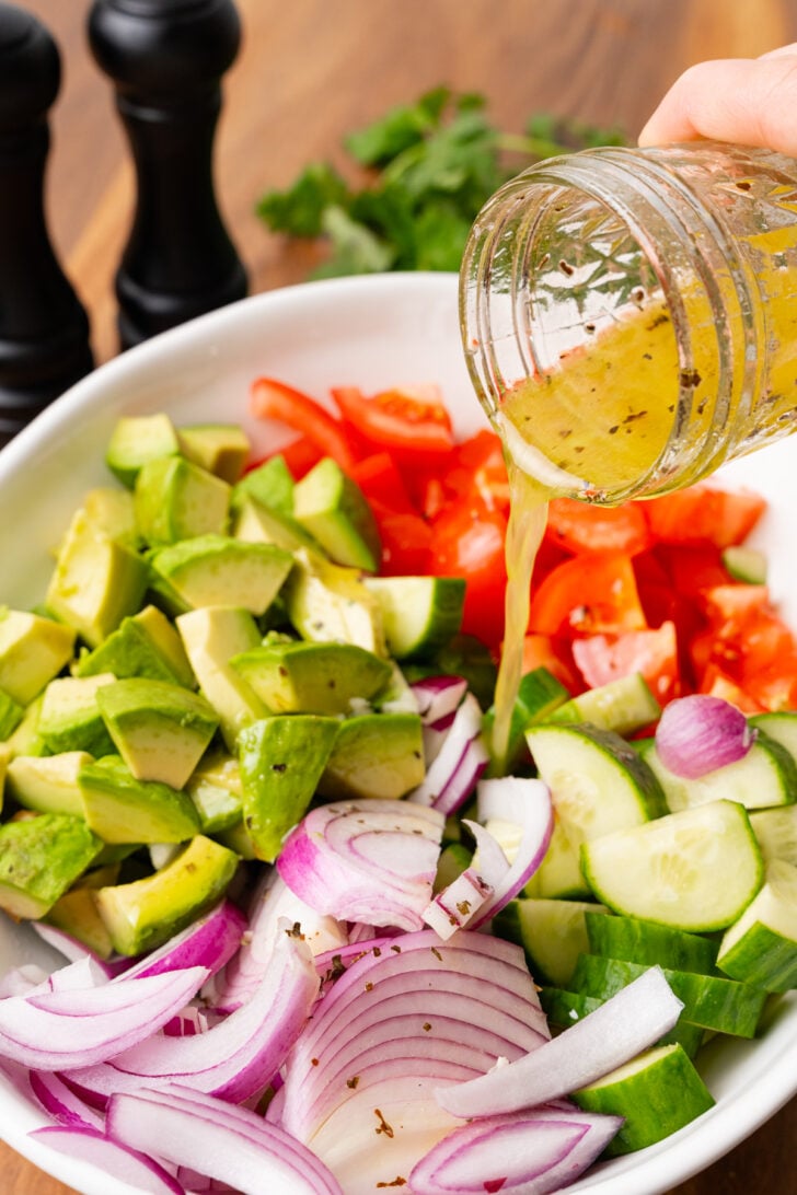 Chopped avocado, cucumbers, tomatoes and sliced onions in a white salad bowl with a jar of homemade salad dressing.