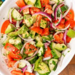 Large white serving bowl with sliced red onions, cucumbers, chopped tomatoes and avocados with homemade dressing and chopped parsley.