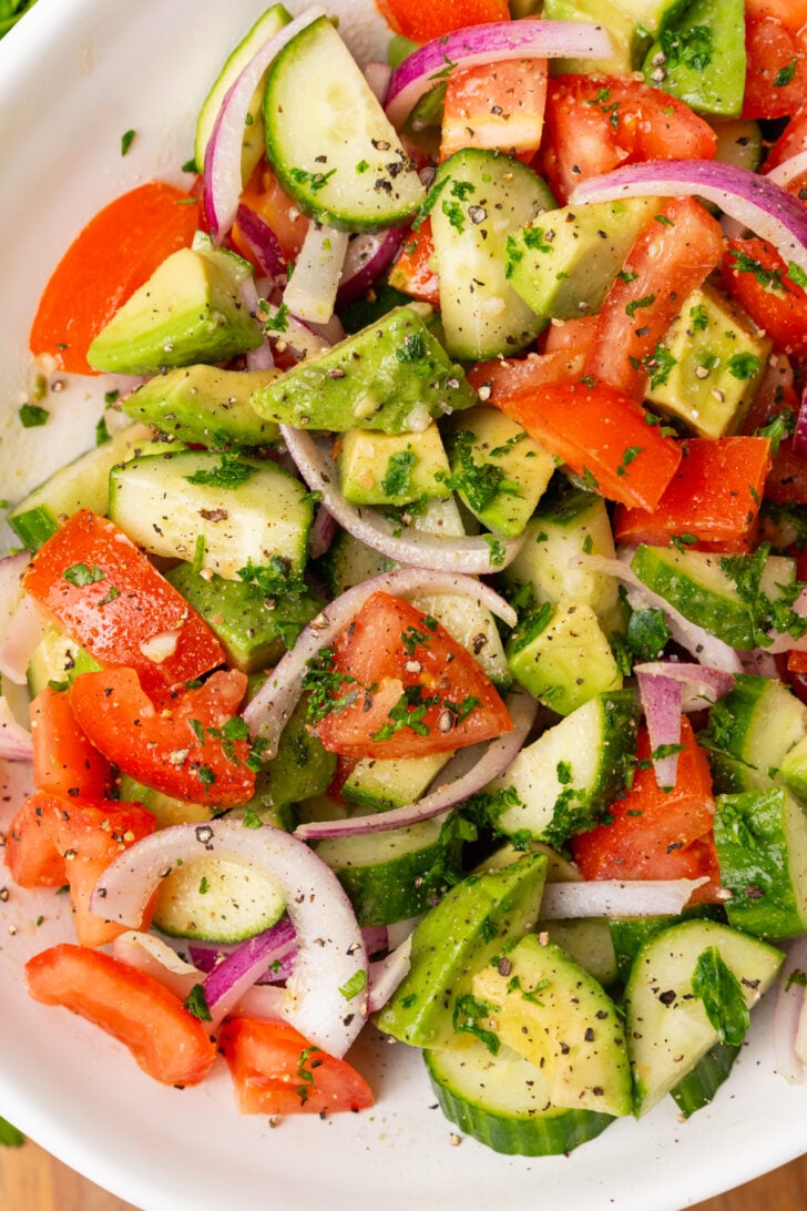 Large white serving bowl with sliced red onions, cucumbers, chopped tomatoes and avocados with homemade dressing and chopped parsley.