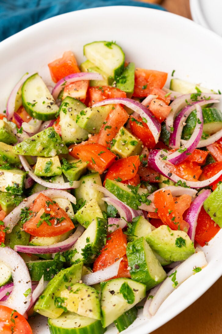 Large white serving bowl with sliced red onions, cucumbers, chopped tomatoes and avocados with homemade dressing and chopped parsley.