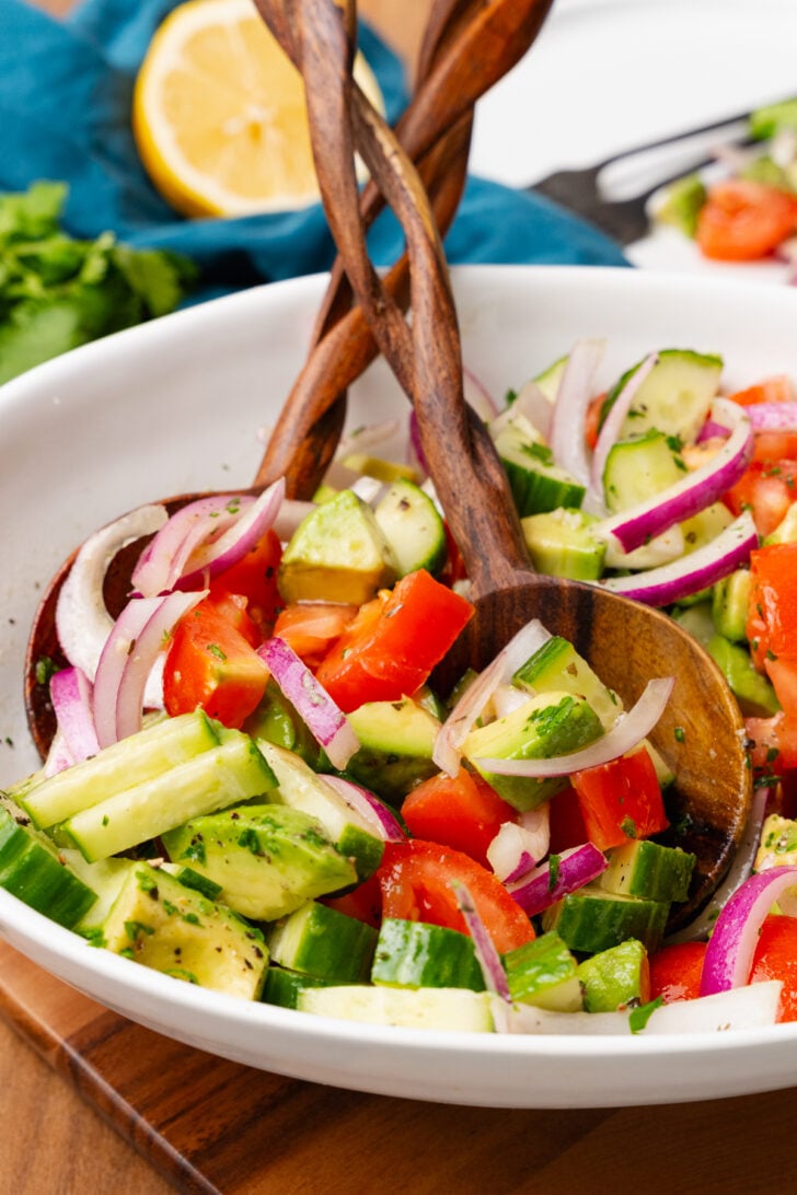 Wooden salad serving spoons in a white bowl with cucumbers, tomatoes and cucumbers.