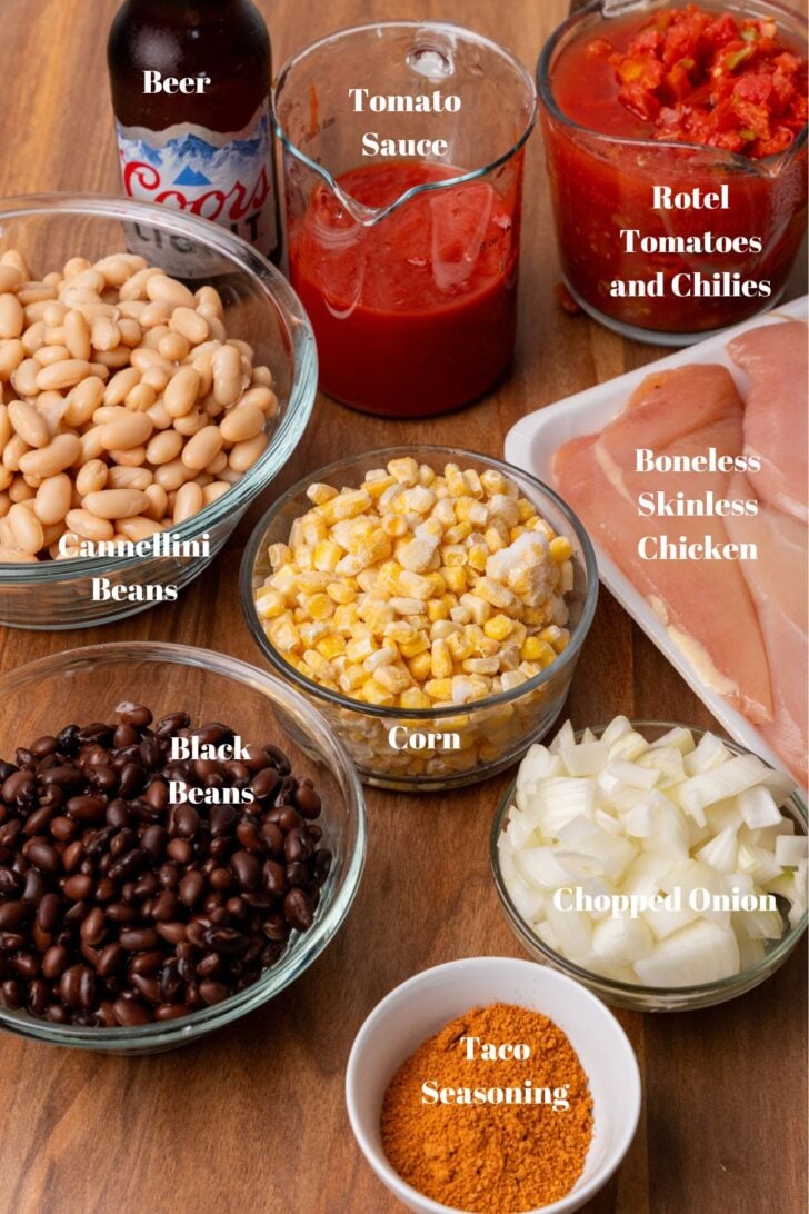 Cannellini beans, black beans, taco seasoning, chopped onions, corn, chicken breasts, rotel tomatoes with chilies, tomato sauce, and beer on a cutting board.