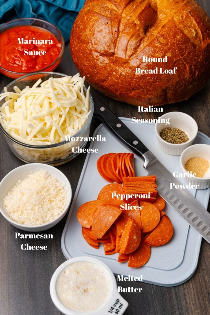 Marinara sauce, round loaf of bread, mozzarella cheese, Parmesan cheese, melted butter, pepperoni slices, Italian seasoning and garlic powder on a white cutting board with a knife.