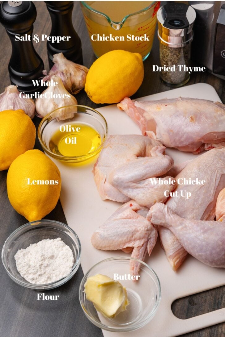 Salt and pepper, chicken stock, dried thyme, whole cut up chicken, garlic, olive oil, lemons, flour and butter on a white cutting board.