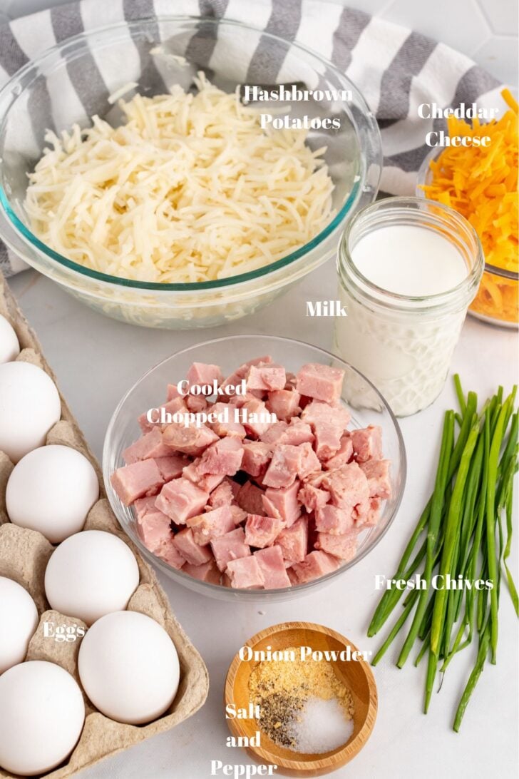 Hashbrown potatoes, cheddar cheese, milk, cooked chopped ham, eggs, fresh chives, onion powder, salt and pepper on a white counter.