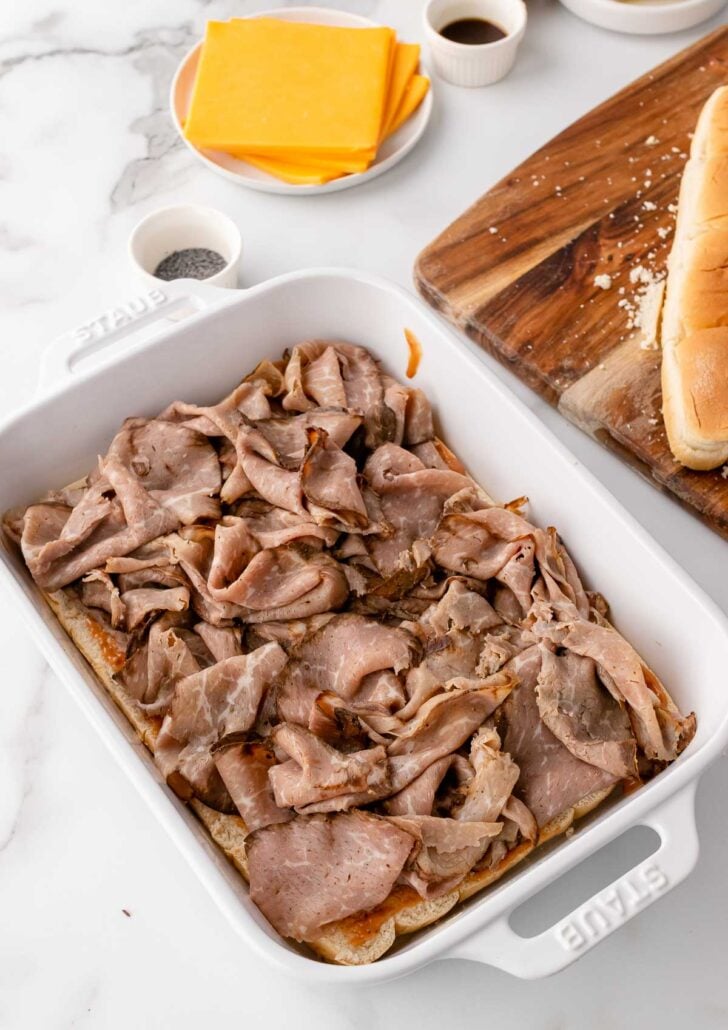 White casserole dish with slider rolls topped with sliced roast beef and a stack of sliced cheddar cheese.