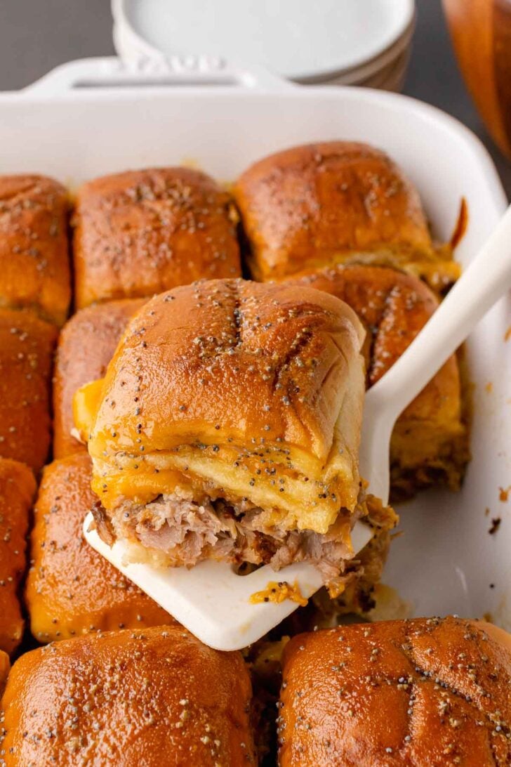 Roast beef slider on a bun topped with poppy seeds baked in a white casserole dish, served with a white spatula.