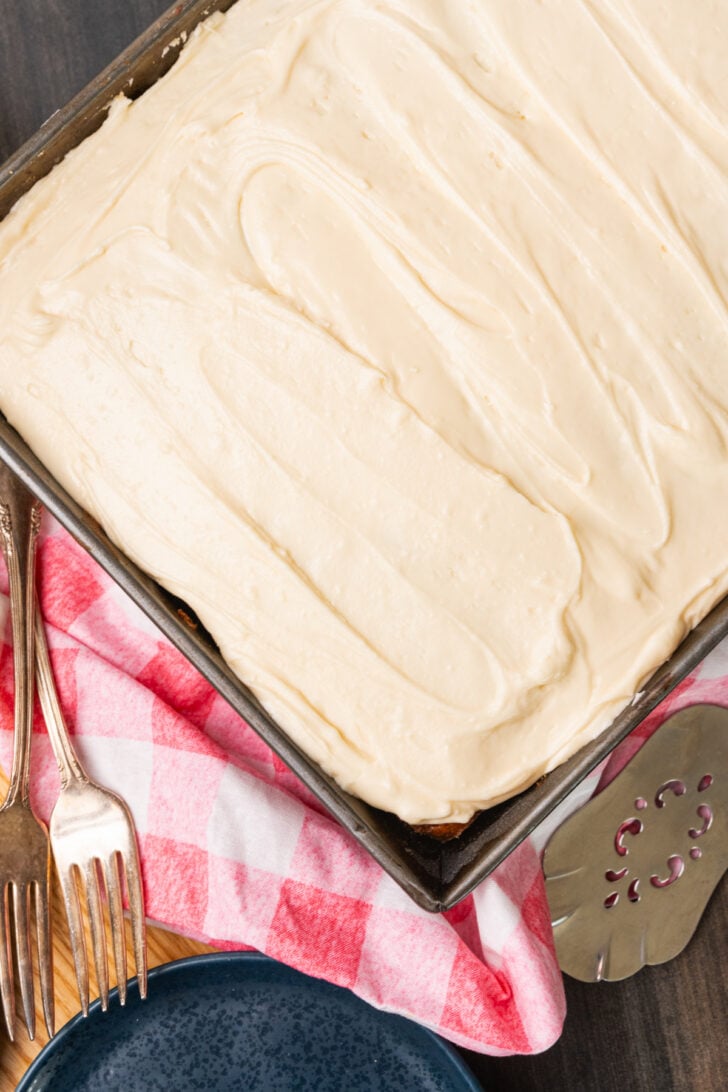 Whole caramel snack cake with creamy white icing in a metal pan cooling on a red and white cloth.