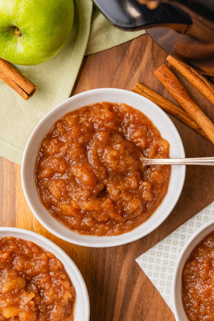 Homemade Crock Pot Applesauce in a white bowl served with a silver spoon on a board with green apples and cinnamon.
