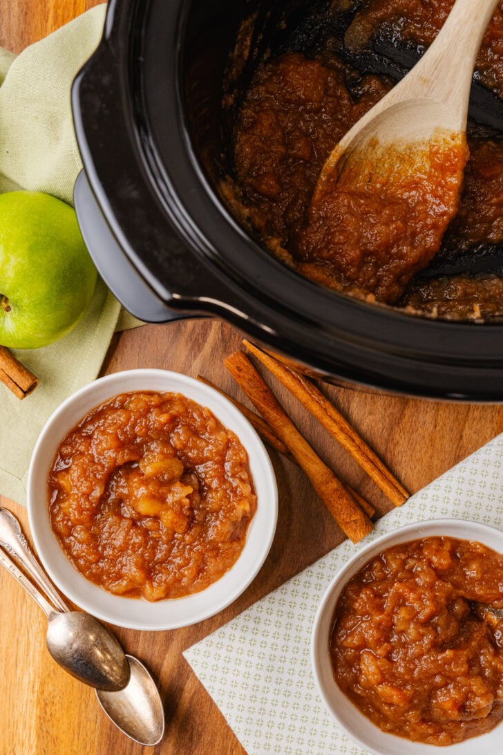 Homemade Crock Pot Applesauce in a white bowl served with a silver spoon on a board with a black crock pot.