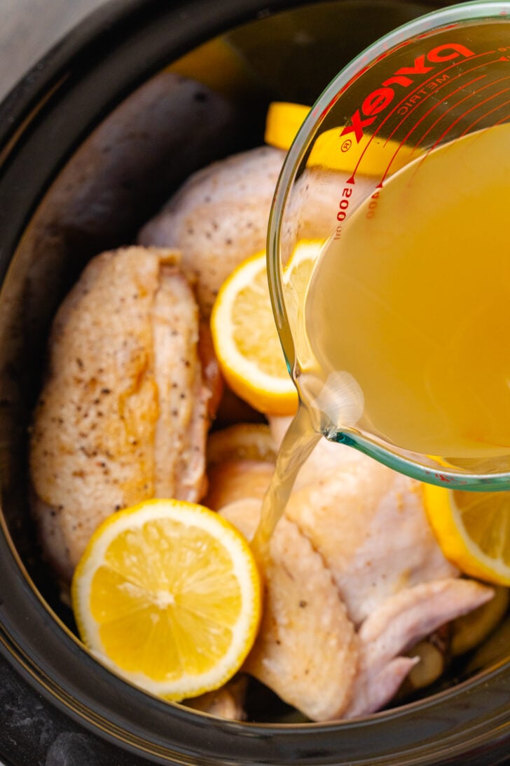 Chicken stock being poured from a glass measuring cup over raw chicken in lemon slices in a black crock pot.