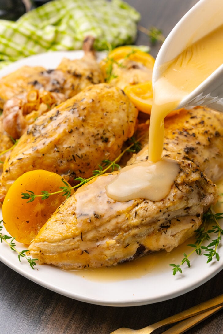 Lemon chicken cooked in the crock pot on a white platter with garlic and a fresh thyme sprigs. and creamy gravy being poured from a white pitcher.