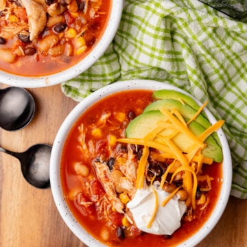 White bowls with taco soup topped with cheddar cheese, sour cream, and avocado on a brown table, with spoons and a green check napkin.