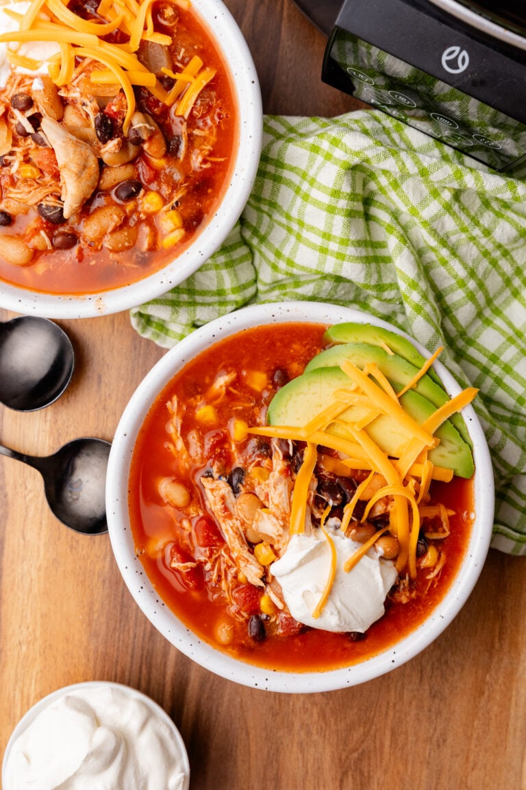 White bowls with taco soup topped with cheddar cheese, sour cream, and avocado on a brown table, with spoons and a green check napkin.