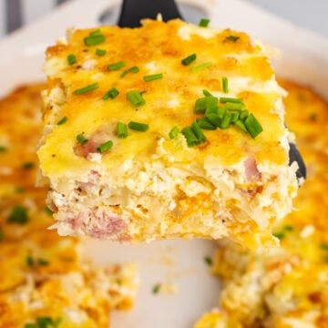 Square slice of ham and potato hashbrown casserole on a black spatula but from a white casserole dish.
