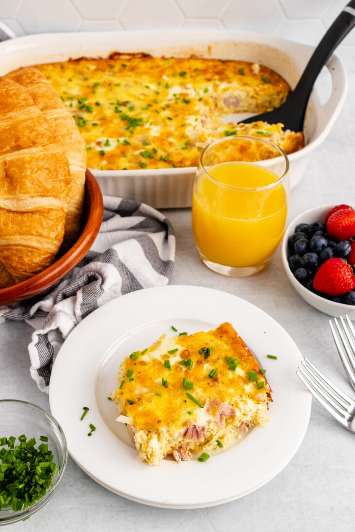 Square slice of ham and potato hashbrown casserole on a white plate cut from a white casserole dish with chopped fresh chives and fresh fruit.