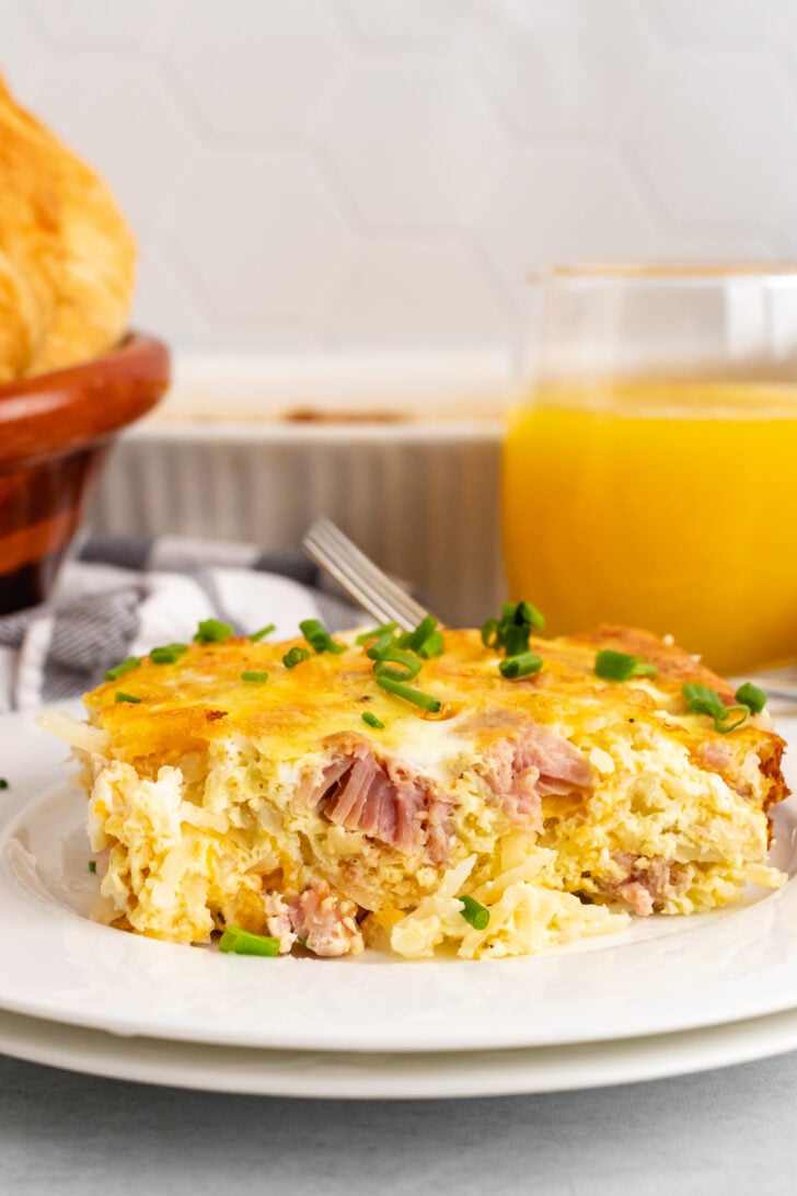 Square slice of ham and potato hashbrown casserole on a white plate with o glass of orange juice.