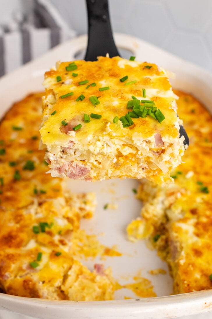 Square slice of ham and potato hashbrown casserole on a black spatula but from a white casserole dish.