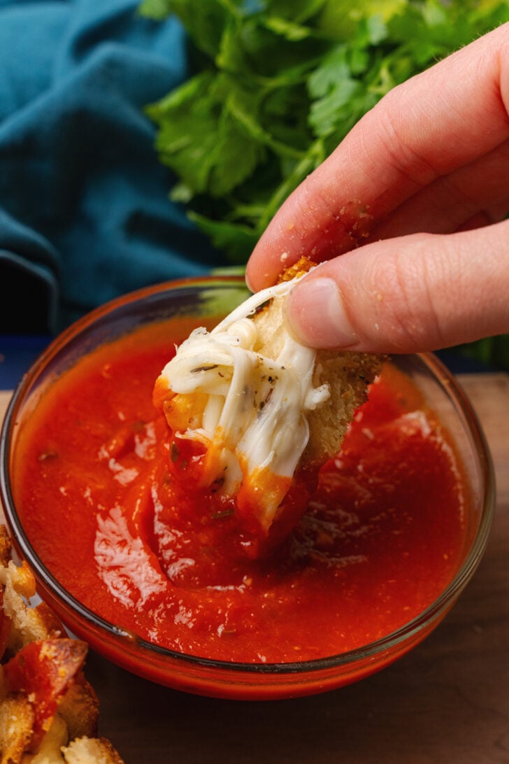 Hand holding a piece of pepperoni pizza bread being dipped into a bowl of marinara sauce.