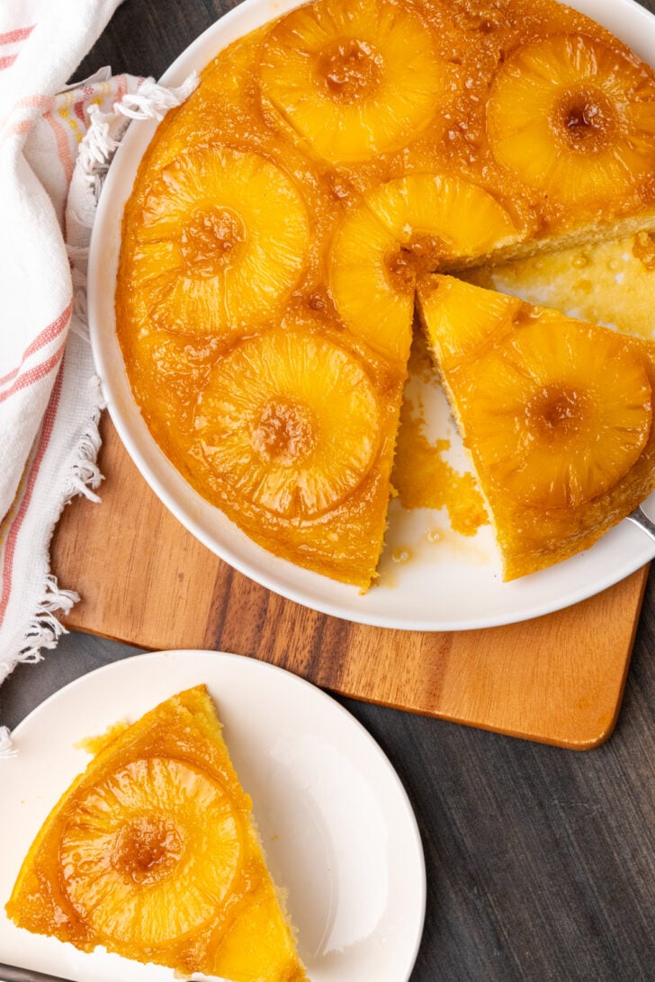 Upside down pineapple cake on a white plate on a wooden board with a slice of cake on the side.