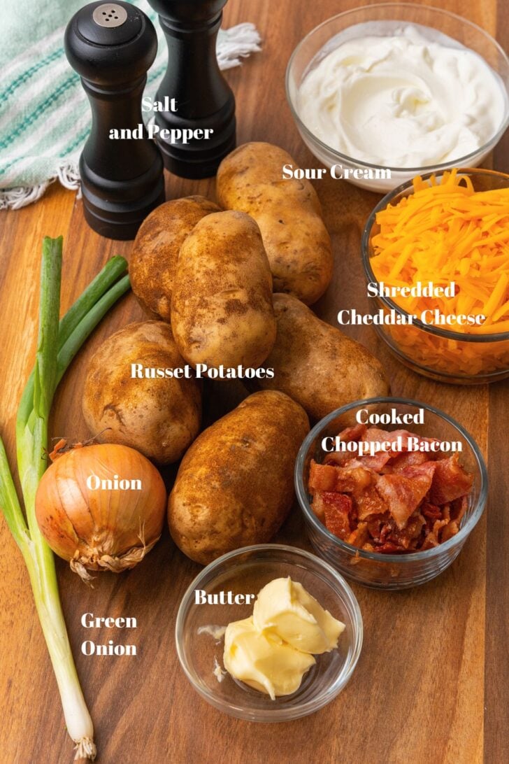 Salt and pepper, sour cream, cheddar cheese, cooked chopped bacon, butter, green onions, onion and russet potatoes on a wooden cutting board.