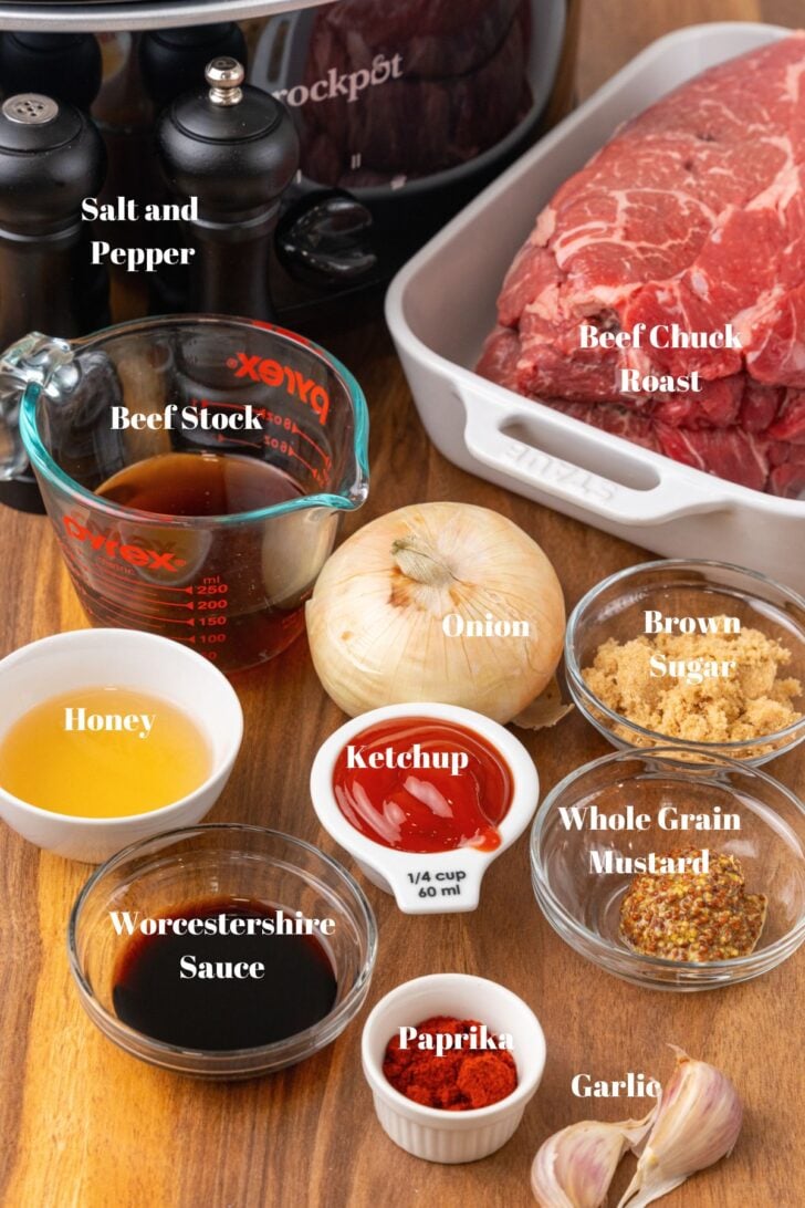 Crock pot, beef chuck roast, beef stock, onion, brown sugar, whole grain mustard, ketchup, honey, Worcestershire sauce, honey, paprika, garlic and slow cooker on a wooden counter.