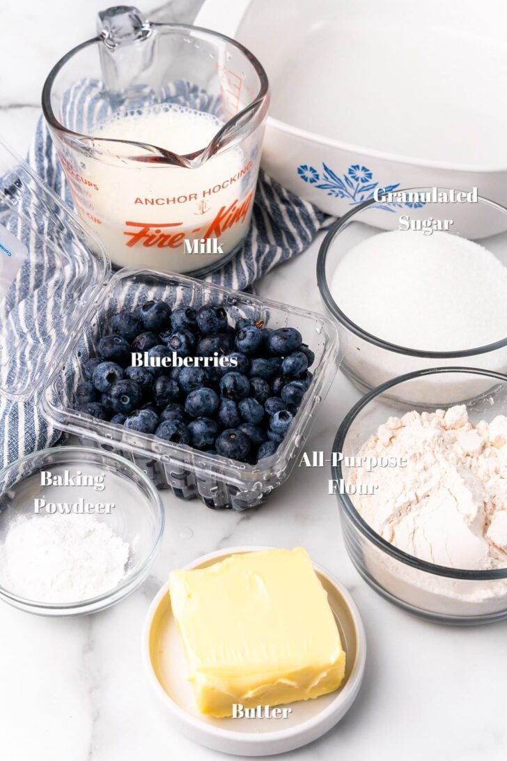 Milk, blueberries, sugar, flour, butter and baking powder in glass containers on a white kitchen counter.