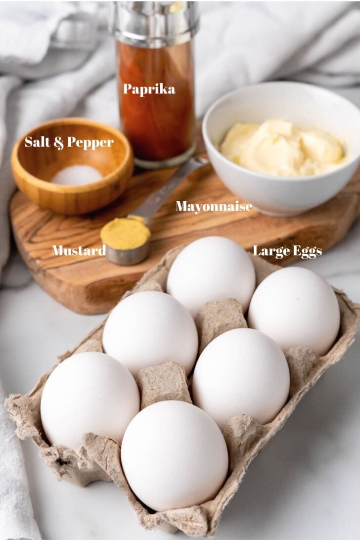 Large eggs, mayonnaise, yellow mustard, salt, pepper and paprika on a wooden board for deviled eggs.