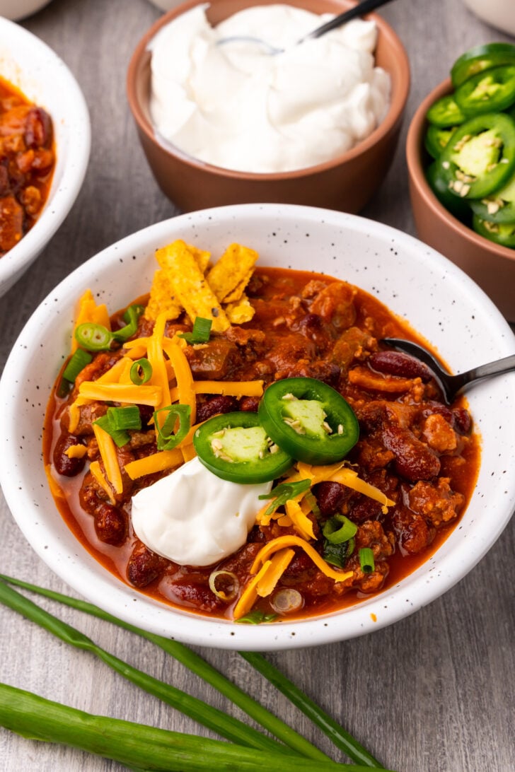 Bowl of ground beef chili in a white bowl served with sour cream, sliced jalapenos and chives served with a spoon.