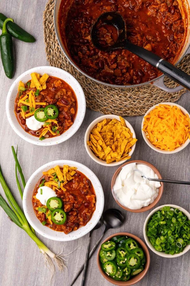 Bowl of ground beef chili in a white bowl served with sour cream, sliced jalapenos and chives served with cheddar cheese and sliced scallions.
