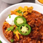 Bowl of ground beef chili in a white bowl served with sour cream, sliced jalapenos and chives served with a spoon.