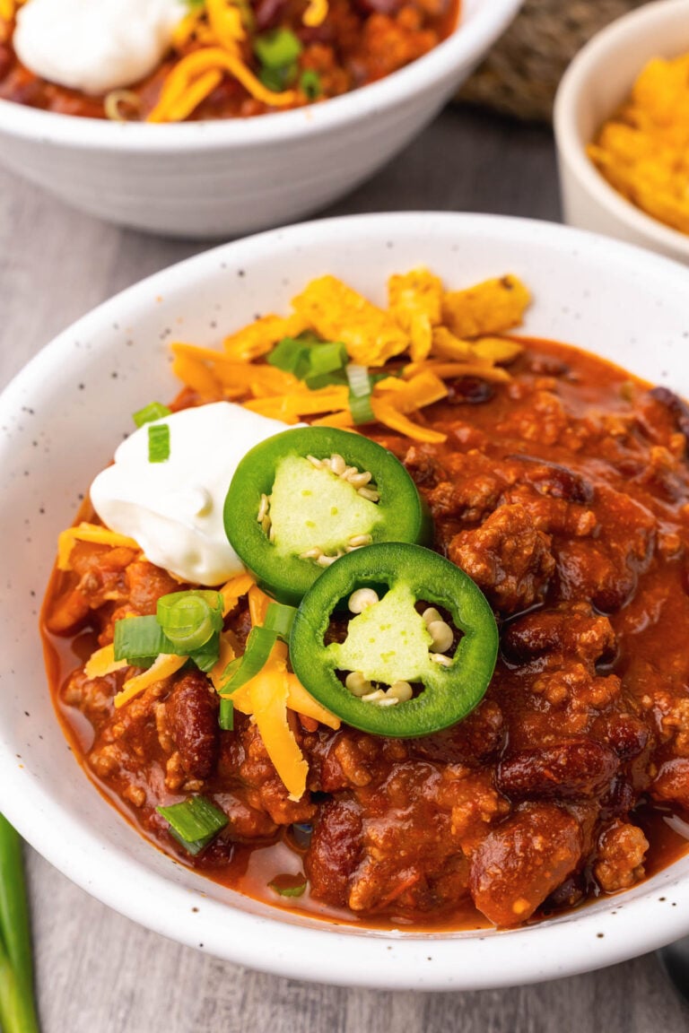 Bowl of ground beef chili in a white bowl served with sour cream, sliced jalapenos and chives served with a spoon.