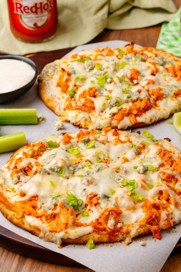 Flatbread topped with buffalo chicken, blue cheese, celery and mozzarella then baked on a wooden cutting board topped with parchment paper.